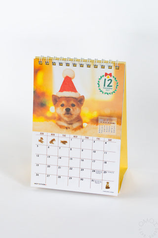 Desk calendar with Shiba Inu in red hat cover, spiral rings, month grid.