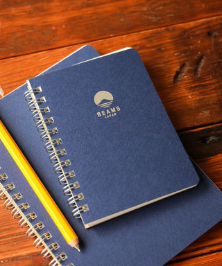 Navy lined notebook with spiral binding and BEAMS Japan logo on cover, beside yellow pencil.