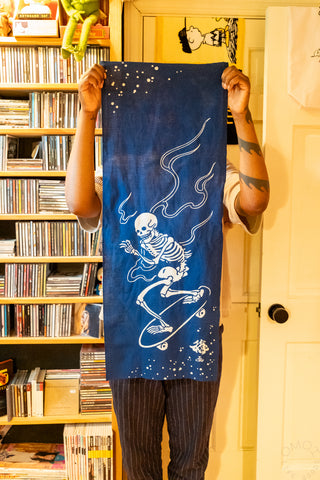 Navy tenugui towel held up, featuring white skeleton on skateboard with wave lines