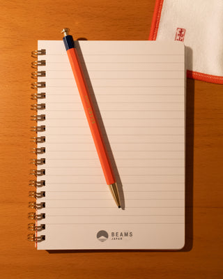 Notebook with an orange pencil on a wooden surface, featuring the Beams brand.