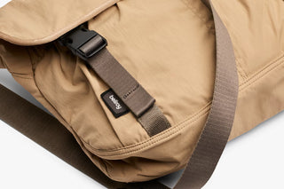 Sand-beige messenger bag with front webbing strap, interior label, and zippered compartment