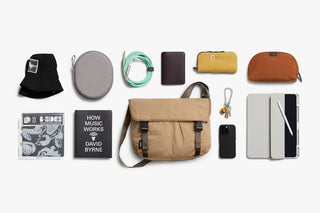 Sand-beige messenger bag shown with accessories: pouch, charging cable, and tech items