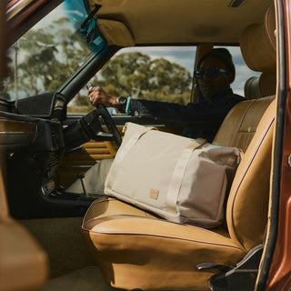 Beige duffel tote bag placed on a car seat, with top handles and front logo patch.