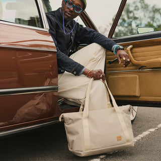 Beige canvas tote bag with top handles and front logo patch, held near a car door.