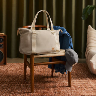 Beige canvas tote bag with top handles and front logo patch, resting on a chair.