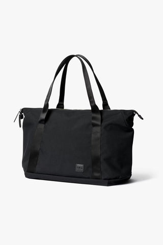 Black duffel tote bag with two top handles, side zip pocket, and front logo patch.