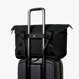 Black duffel tote bag with trolley strap and top handles, shown attached to luggage.