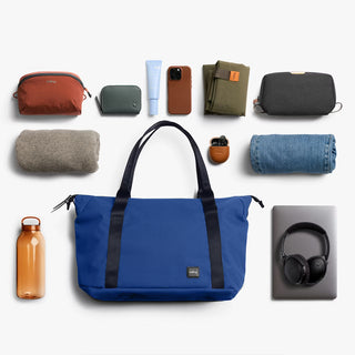 Indigo travel tote bag with accessories shown nearby, black straps, and front logo patch