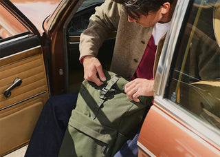 Olive green rolltop backpack on a person, adjusting shoulder strap near car door