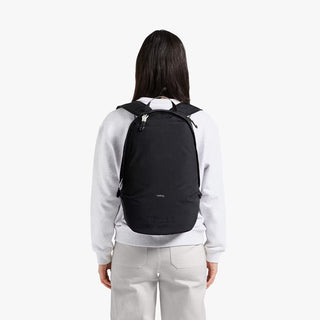 Black daypack on a person, showing top handle and padded shoulder straps from the back