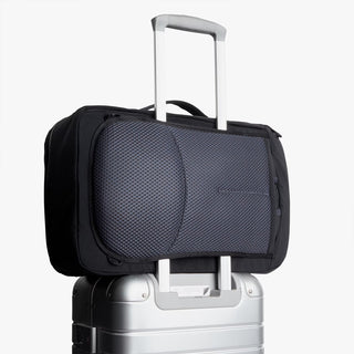 Black travel backpack with padded front panel and top handle, mounted on luggage handle