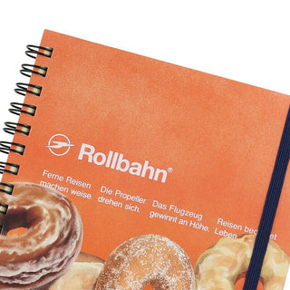 Large spiral notebook close-up, orange cover with Rollbahn logo and donut illustrations