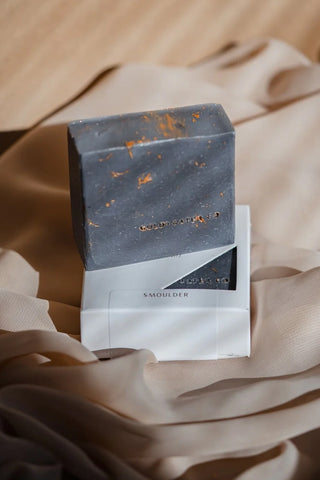 Handcrafted soap bar, dark blue-gray with speckled texture, wrapped in white paper.