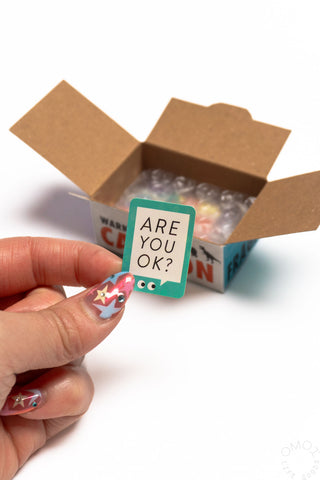 Hand holding ARE YOU OK? sticker with open sticker seal box behind