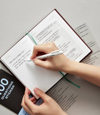 Open diary notebook with printed pages, hands writing with a pen and green ribbon