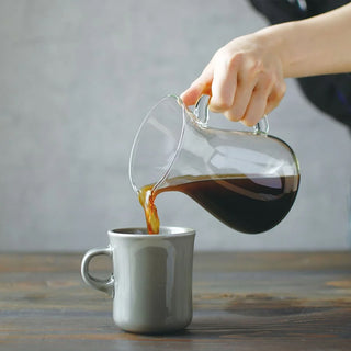 Hand pouring dark coffee from glass carafe into a white ceramic cup