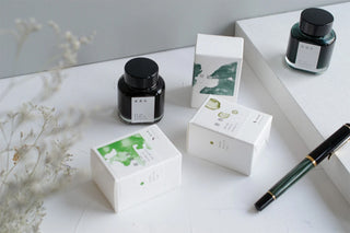 Set of black glass ink bottles with white gift boxes and a green pen.