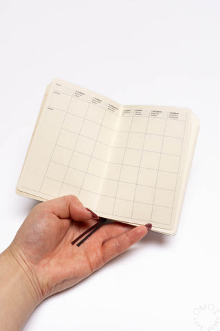Pocket log diary open showing weekly grid layout on cream pages; hand holding