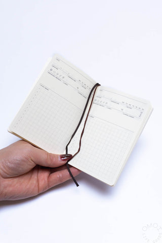 Pocket log diary open with two cream pages and dark ribbon tie; hand holding