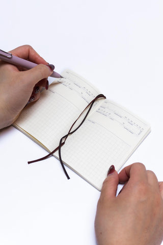 Pocket log diary open with dark ribbon bookmark and checklist grid; both hands visible