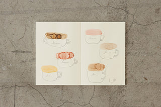 Thick cream notebook open to pages with cute coffee cup sketches in colors.
