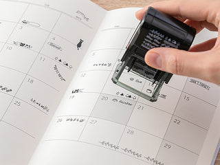 Hand holding a black stamp over a printed planner with small illustrated icons.