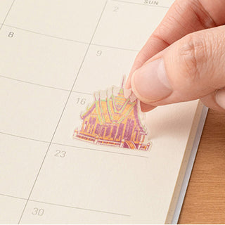 Hand placing a colorful Thai illustration sticker on a dated planner page