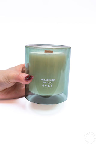Green glass jar candle with white wax and wood wick, held in a hand.