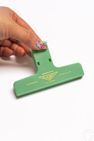 Green plastic binder clip with PENCO Clampy PLA-clip logo, held by hand