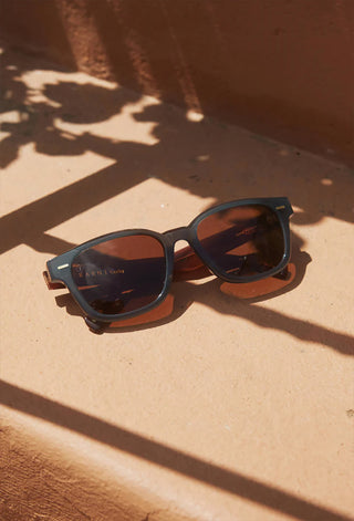 Dark sunglasses with teal inner frame and dark brown lenses on a sunlit surface