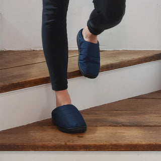 Person wearing navy outdoor slippers while standing on a wooden floor