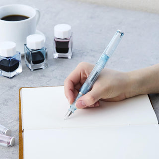 Glassy fountain pen with light-blue transparent body and metal nib, held writing on paper