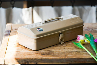 Beige metal tool box with latching lid on wooden table, ready for tools