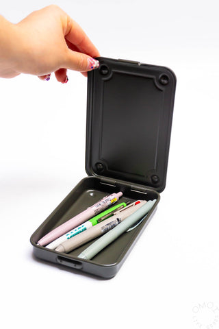 Olive green trunk-shaped toolbox opened by hand, with pens and pencils inside