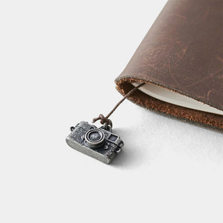 Pewter camera charm pendant on dark cord, near brown leather notebook edge