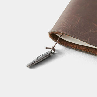 Pewter pen charm dangling from dark cord beside a brown leather notebook cover