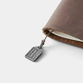 Pewter luggage charm tag on brown leather strap near a stitched notebook edge