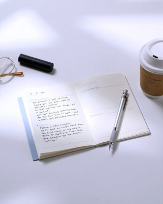 Open A6 journal pages with handwritten notes, pen, and small coffee cup on desk