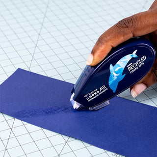 Hand holding MONO Air tape applicator dispensing adhesive onto blue paper