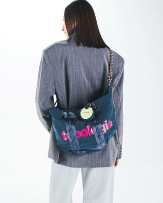 Deep teal duffle bag worn crossbody with round tag and logo print
