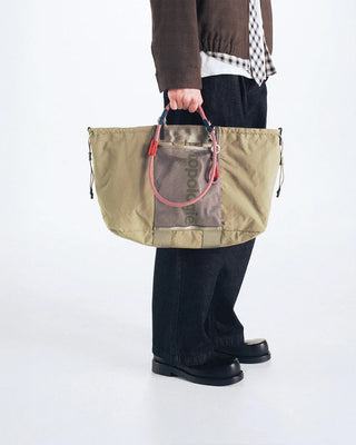 Khaki tote bag with top handles and colorful trim; shown being carried on a person.