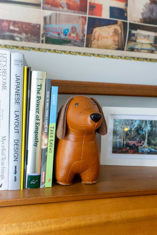 Tan dachshund bookend on a wooden shelf beside books and decor