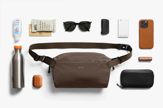 Brown fabric sling bag with adjustable strap, centered among travel accessories.