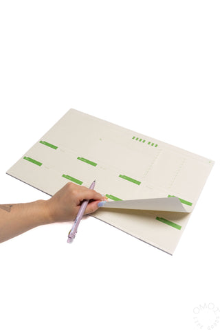 Two-week desk planner with green day blocks, open with pen writing