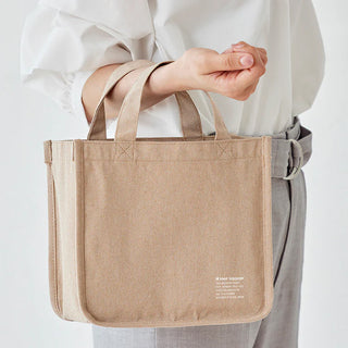 Beige canvas tool tote held by handles with reinforced edges and stitched front pocket panel.