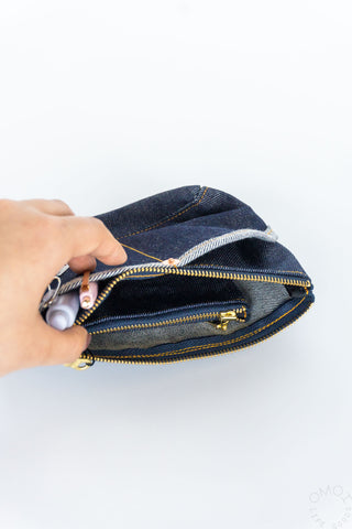 Hand holding open denim pouch; gold zipper, inner divider, keyring clip visible
