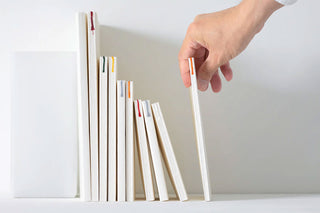 Off-white and plain MD PAPER notebooks of different sizes are lined up on a bookshelf of similar color. Different colored bookmark strings can be seen from the face-out spines. A light skin person's hand is picking up one of the notebooks.