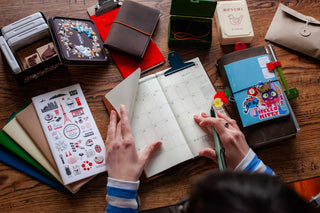 In the center is N's TRAVELER'S notebook opened to a monthly calendar as they are about to write. Surrounding the notebook are other TRAVELER'S notebook inserts, a metal tin full of stamps, more TRAVELER'S notebooks, and stationery containers holding TRAVELER'S band accessories. 