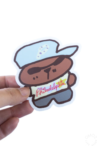 Hand-held die-cut mascot sticker: brown character, blue hat, red Philadelphia text