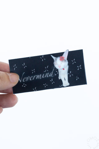 Vinyl sticker held in hand: black rectangle with white cat “Nevermind” text and sparkles